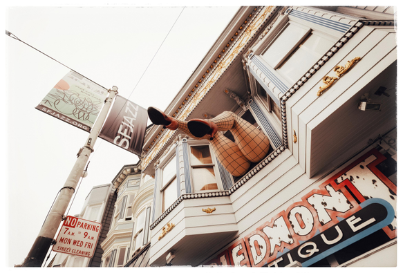 A picture taken on a foggy day on Haight Street in San Francisco... looking up at windows above a storefront on that busy street. Hanging out of one of the windows on the building's second story: two giant fiberglass legs in fishnet stockings and red high heels. The legs are enjoying the breeze.