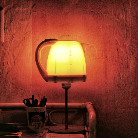 Das quadratische Foto zeigt eine ungewöhnliche, improvisierte Tischlampe, deren Lampenschirm aus einem leuchtenden, gelb-weißen Wasserkocher aus Kunststoff besteht. Die gesamte Szene ist in ein warmes, rötlich-oranges Licht getaucht. Der Wasserkocher ist mittig auf dem schlanken Metallgestell einer Stehlampe montiert und strahlt von innen heraus so hell, dass seine Füllmengenmarkierungen auf der Vorderseite deutlich zu erkennen sind. Der Griff des Kochers zeigt nach links, während der Ausguss nach rechts weist. Die Lampe steht auf einem rustikalen Holztisch oder einer Kommode direkt vor einer grob verputzten, unebenen Wand, die durch das Licht tiefe Schatten und eine kräftige rote Färbung erhält. Links neben dem Lampenfuß steht ein gemusterter Becher, der als Stifthalter dient und mit mehreren dunklen Stiften gefüllt ist. Im Vordergrund sind zudem vage die Kanten eines Notizblocks und einiger Papiere zu sehen, während ganz oben links im Anschnitt schwach ein Lichtschalter oder eine Steckdose an der Wand ausgemacht werden kann.