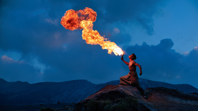Side shot of a man breathing fire against a blue hour mountainous backdrop