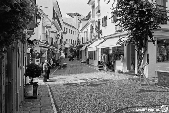 A quaint cobblestone street in Marbella Old Town  lined with shops and whitewashed buildings, featuring people engaged in conversation and vibrant greenery. The scene captures a charming, relaxed atmosphere typical of a Mediterranean village.