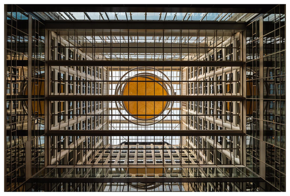 Blick von unten nach oben in eine Art Lichthof. Um den Hof herum sind zahlreiche Fenster sichtbar. Mittig im Lichthof hängt ein rundes gelb-orangefarbenes Objekt.