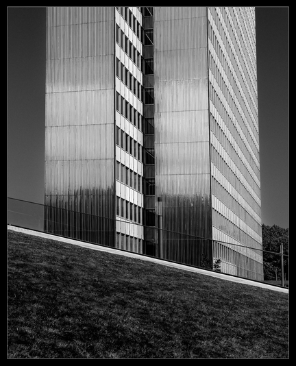 Hinter einer begrünten Rampe (schräge Grasfläche/Dach) steht ein silbrig schimmerndes Hochhaus.