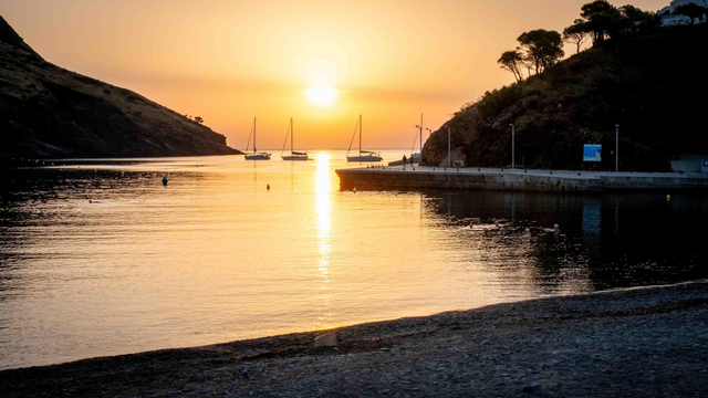 Badia de Portbou amb velers ancorats i l'embarcador a la dreta. Sol mig ennuvolat amb un reflex taronja sobre les aigües de la mar.