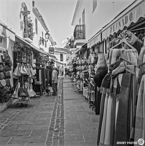A narrow street lined with shops displaying bags, textiles, and souvenirs. Two people are engaged in conversation, while a small dog stands nearby. The scene is captured in black and white, enhancing the vintage market atmosphere.