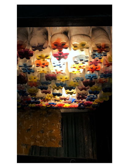 The dark interior of the entryway to Hidden Gem Cafe in Hanoi, Vietnam. Yellow paint flakes off the walls, which are also partly patched up with corrugated metal sheets. Looking up, we see that the ceiling is completely made up of old water bottles, tightly packed in a vertical orientation so that we see their star-shaped bottoms which are painted in different colors. The ceiling acts as a skylight, with daylight shining through some of the bottles.