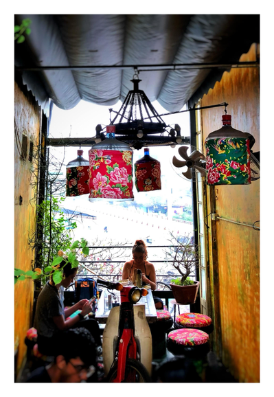 The second floor of the Hidden Gem Cafe. Customers sit by a floor-to-ceiling window overlooking the street below, which is overexposed in bright sunlight. The seating in this area is made of repurposed rubber tires topped with round patterned cushions.  There’s also the front section of a motor scooter with its handlebars, headlight and wheel, repurposed as a seat back. Overhead is a chandelier made of large water cooler jugs wrapped in colorful floral fabrics.