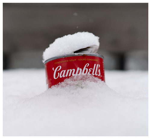 The upper half of an open can of Campbell's soup ensconced in a bed of freshly-fallen snow in front of a dark background. On the top of the can, a cap of snow. The photo is reminiscent of a certain pop art classic...