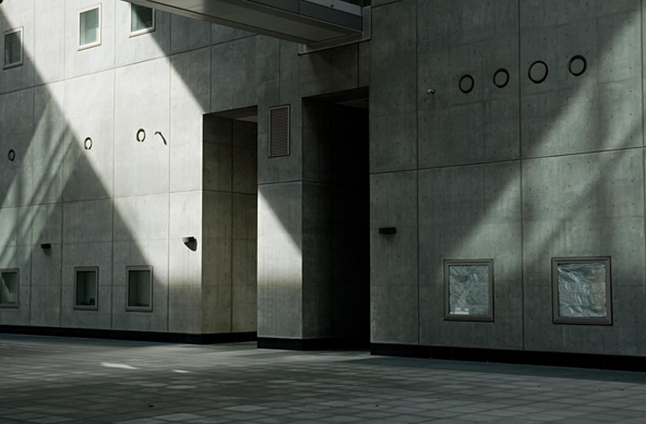 A photo of a facade of a modern building with some, minimalist concrete design, featuring square and circular cutouts. Light rays and shadows fall diagonally across the wall and the tiled floor.