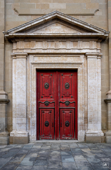 Color photo of a large red double door with square fluted stone columns on either side and topped by classical architrave and triangular pediment.