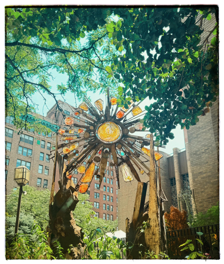 A color photo of a large sun made out of welded iron pieces and fitted yellow and orange pieces of glass. In the center of the sculpture is a large round yellow piece of glass attached with some wire to an outside piece of metal. Protruding from that are many individual metal spikes. The different length spikes have different shaped orange and yellow pieces of glass wedged in between them and attached by wire. It is being held up by two pieces of metal on either side that almost look like chunks of rock. To the right side is a little bit of the light brickwork from the front of the church. On the left side is a street lamp with a basic brick building behind it. There's lots of greenery hanging over the top of the photo with various leaves from different types of trees. A little blue sky is poking through.