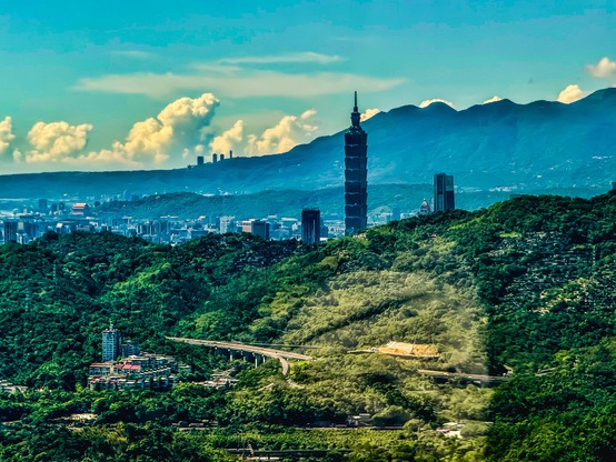 台北市景观，台北101大厦矗立在绿色山丘之间，背景为蓝天白云和远山。
