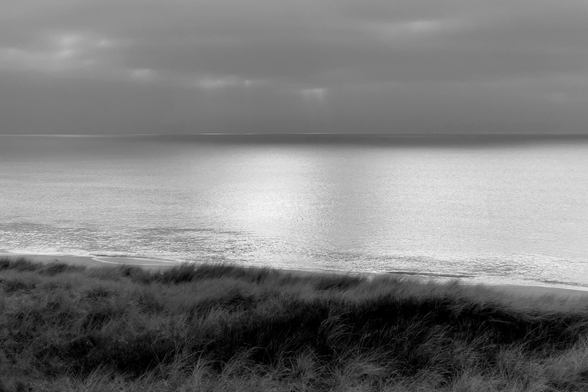 Eine schwarz-weiße Aufnahme vom Meer: Im Vordergrund Dünen mit Gras, dahinter die Nordsee, die silbrig glänzt, am Horizont ein heller Streifen und darüber ein dunkelgrauer wolkenverhangener Himmel. Die Stimmung ist friedlich.

A black-and-white photograph of the sea: in the foreground, grassy dunes, behind them, the North Sea, glistening silver. On the horizon a pale strip and above it, a dark grey, cloud-covered sky. The atmosphere is peaceful.