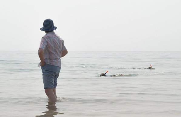 A photo looking out on a calm sea under an overcast sky, showing a person wearing a shirt, shorts, and a wide-brimmed hat standing in shallow water at the edge of the beach. In the water, two swimmers with snorkels are visible, partially submerged. 