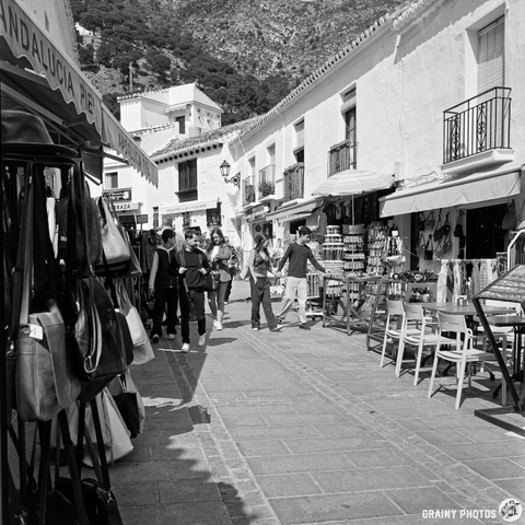 A bustling Street in Mijas with souvenir shops selling textiles and crafts and street cafes. People walk among the stalls, enjoying the vibrant atmosphere in a picturesque town surrounded by mountains. The scene is captured in black and white, adding a timeless feel.