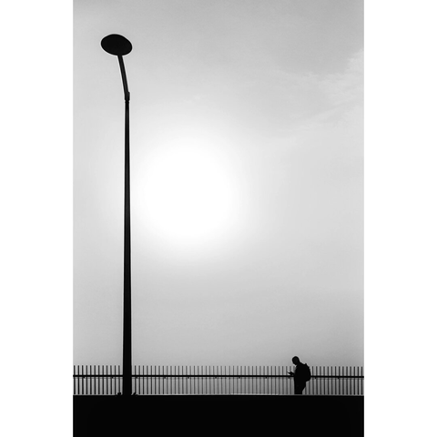 Minimalistic street photography taken in Luxembourg city showing the silhouette of a man walking on a bridge next to a lamp post. The photography faces the sun, appearing in the fog as a white halo.