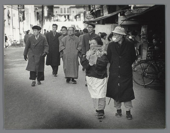 The image depicts a street scene from what appears to be mid-20th century Japan, given the style of clothing and overall atmosphere. A group of people is walking down an urban street; their attire suggests it might be cool weather as most are wearing coats or overcoats with hats for headwear.

In the foreground, there's a man dressed in dark clothes holding what seems to be a child wrapped in cloth on his back, indicating that he could be traveling with family. The second individual walking beside him is older and wears glasses; they have a mask around their chin instead of covering their nose and mouth completely, possibly due to the temperature or local custom at the time.

The background includes other individuals wearing coats who are also in motion on the street. Some buildings can be seen along with what looks like market stalls or shops lining one side of the road. A bicycle is being carried by a person further back from the camera, adding to the sense of movement and daily life captured here. The overall black-and-white tone gives it an aged feel.

The photograph captures a slice-of-life moment that provides insight into social norms and fashion in Japanese urban environments during its era.