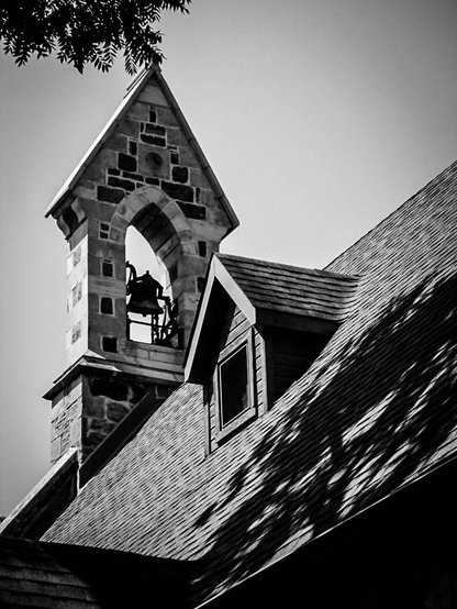 Looking up towards the roof and bell tower on a small church. A bell can be seen in the brick tower and a small dormer window is also seen. Shadows from a nearby tree are seen and a tree branch in the top left of this black and white image. 