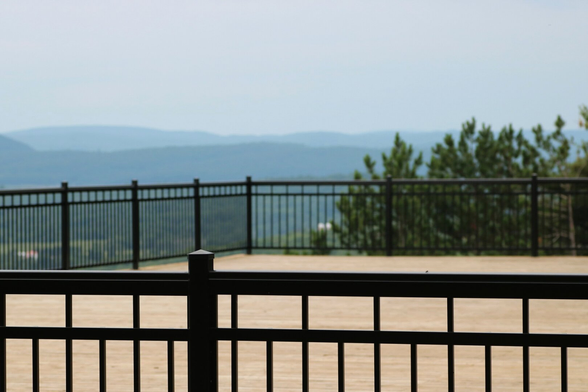 Photograph of a large, light-colored wooden patio enclosed by a black iron fence with straight bars, built on a promontory on the edge of a mountain. In the foreground, a fly is perched on top of the black railing. In the background, the landscape consists of bluish mountains under a gray sky, green trees, and farm buildings visible between the bars.

Photographie d'un grand patio en bois blond entouré d'une clôture noire en fer, avec des barreaux droits, construit en promontoire sur le rebord d'une montagne. En avant-plan, sur le dessus de la rampe noire, une mouche est posée. En arrière-plan, le paysage fait de montagnes bleutées sous un ciel gris, des arbres verts et des bâtiments de fermes visibles entre les barreaux.