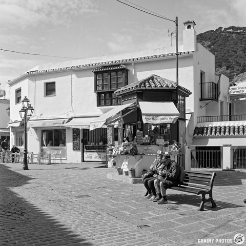 A black-and-white photograph of a quaint street scene featuring a street stall. Two people sit on a bench in front of the shop, enjoying the atmosphere, with a scenic hillside in the background. Historic charm and a relaxed vibe are evident.