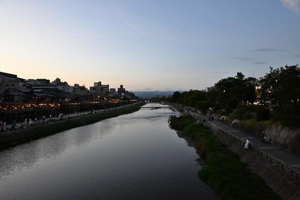 時間帯は日の入り直後くらいだけど、西の空はやや明るい。鴨川の西側は納涼床の明かりと河原に座る人影が奥(北側=三条通方面)に向かって並んでいる。/ 2026.07.26 四条大橋から