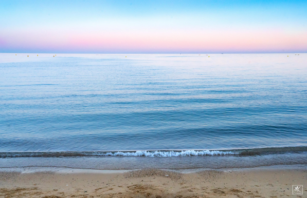Color photo looking out to the horizon from a beach on the coast of Catalonia with small waves approaching the shore and a colorful post-sunset sky above.