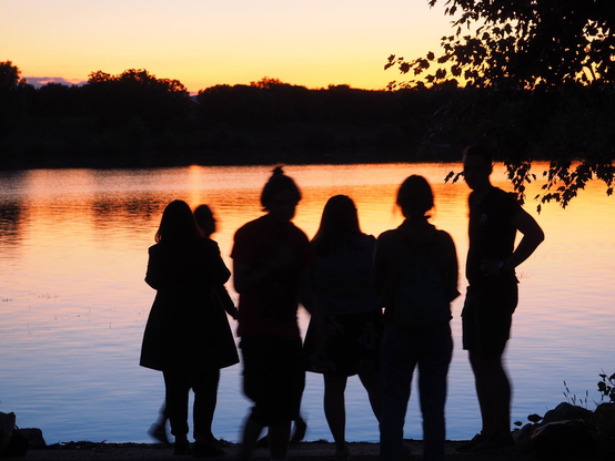 6 Personen als schwarze Umrisse vor einem Fluss in der Abendsonne, Das Wasser hat einen Farbverlauf von oben orange zu unten blau. Am Horizont teilt eine schwarze Baumreihe das Wasser vom Himmel. 