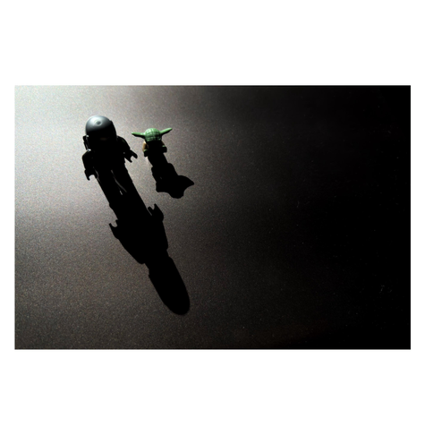LEGO figures of the Star Wars characters Din Djarin (the Mandalorian) and Grogu (a.k.a. Baby Yoda) standing on a dusty metallic surface, backlit so that they’re mostly in silhouette and casting long shadows in front of them. The scene is viewed from above and at an angle so that the toys are standing at the upper left and their shadows stretch diagonally towards the center of the image. The bright light source behind them softens into darkness as it spreads to the edges and far corners of the photo.