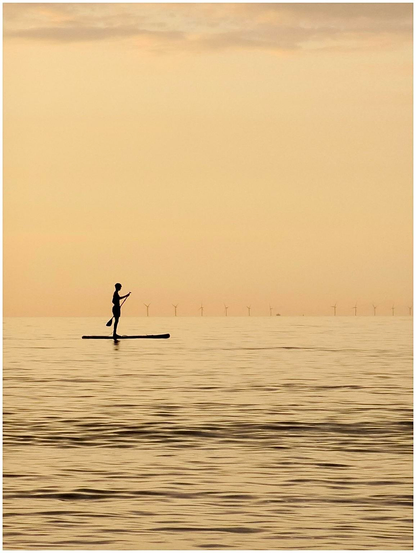 A silhouette of a person paddleboarding on calm water during sunset, with wind turbines visible in the background. The scene features warm orange and yellow tones in the sky.