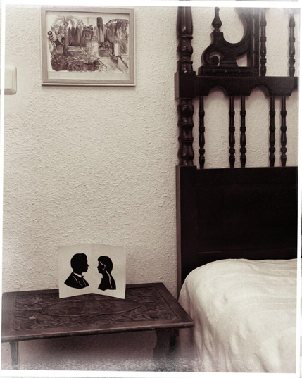 Ausschnitt eines Zimmers, rechts ein Bett mit katalanischen Kopfteil, links daneben ein Nachtisch. eine Klappkarte mit zwei schwarz-weiss aus schwarzem Karton geschnittenen Profilen sehen sich an. Darüber ein Aquarell-