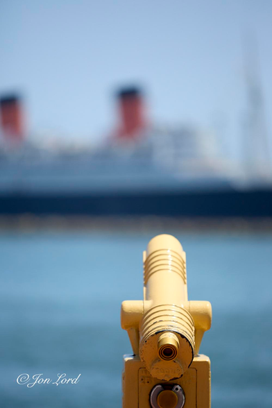 This is a colour abstract photo in portrait format of a waterfront telescope (as in deposit a coin and watch for a minute or two) in the foreground, a body of water in the middle ground and a moored ocean liner in the background. Long Beach, California (2013).

In the foreground and slightly to the right of centre is the bright yellow and heavily built telescope. The telescope is viewed from behind and slightly above with the camera being in line with the body of the telescope and with it filling half the height of the photo. Only the upper portion is visible with the box like device to collect the money resting on the base of the photo and with the cylindrical, almost cannon like, telescope mounted on a gimbal on top and raised upwards ever so slightly towards the sky. Beyond the telescope is the body of medium blue, almost turquoise, water: rising from the base of the photo upwards to the half way up level in the image. In the background and blurred out of focus is a large ocean liner with just part of the forward section of the ship in view (the bow is out of view to the right). The ship is viewed in profile, is facing to the right, is filling the entire width of the photo and is about one km away. The ship's hull is black, the superstructure is white and has two large cylindrical red funnels with a black stripe on the top. Above is a light blue, clear and cloudless sky.

The ships is the RMS Queen Mary, retired since 1967 to Long Beach, California and repurposed as a hotel and museum.