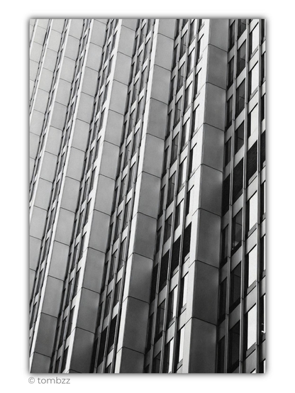 A black-and-white architectural photograph showing a tightly framed section of a modern building facade. Repeating vertical bands, rows of narrow windows and projecting modules create a raw, almost mechanical rhythm. The frame is slightly tilted, causing the regular lines to run diagonally and giving the static structure a distinct sense of movement. Light exterior panels contrast with dark windows, recessed sections and rectangular ventilation grilles. Soft light shapes the successive folds of the façade and emphasises their depth, while deeper shadows separate the individual segments. With no sky, street or clear point of reference, the building loses its scale and begins to resemble an abstract composition made of lines, rectangles, light and shadow. The image is defined by cool geometry, order and a consistent repetition of forms.
