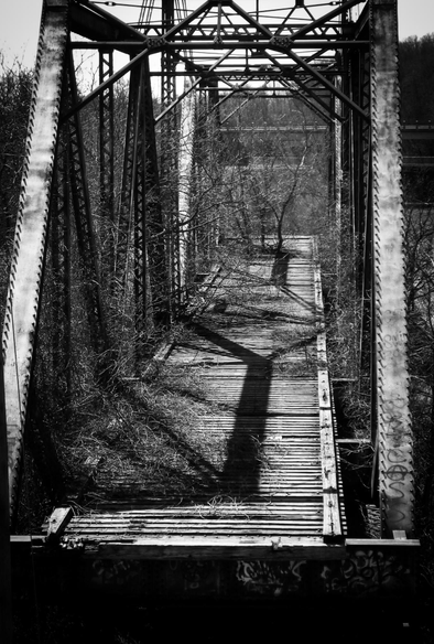 Looking down upon a old disused train swing bridge with the train rails removed from it. The tall bridge like steel structure is seen as well a shadows from the bridge itself as well as nearby large trees. This is a high contrast black and white image. 