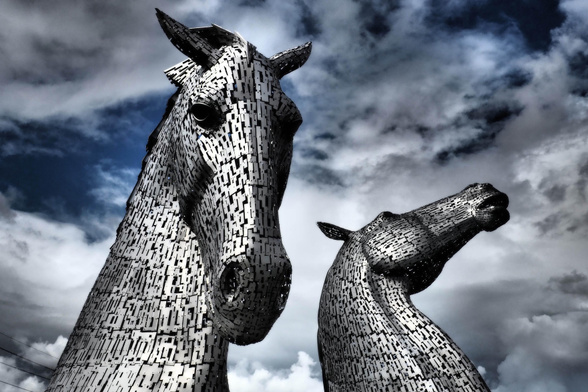Zwei Pferdeköpfe aus Stahl, jeweils rund 30 Meter hoch, vor einem dramatisch bewölkten Himmel, aus der Untersicht fotografiert. Die Skulpturen sind von Andy Scott und stehen in Falkirk, Schottland.

Two steel horse heads, each around 30 metres high, set against a dramatically overcast sky, photographed from below. The sculptures are by Andy Scott and are located in Falkirk, Scotland.