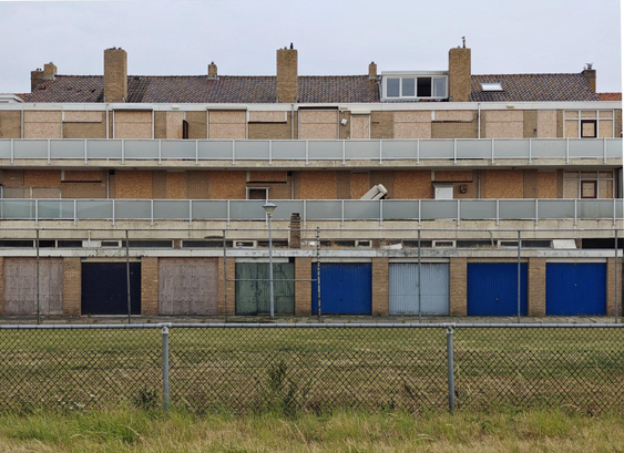 A photo of a three-story brick building: the ground floor has blue garage doors, while the upper floors show many boarded-up windows and doors. On the top floor a single set if windows is still clear, with one window open.