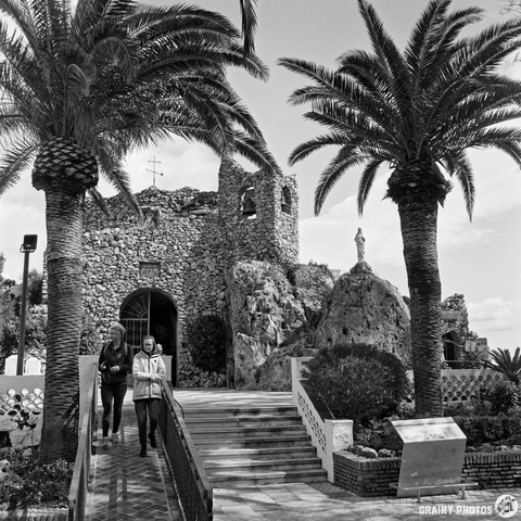 Ermita de la Virgen de la Peña with a statue and a bell tower in the background. Two people walk down a ramp lined with palm trees. The scene is captured in black and white, highlighting the architectural details and natural surroundings.