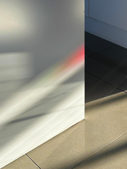 Sunlight, through a window with red and green stained glass panels, brings light, colour, and shadows to otherwise plain white kitchen cabinets and a grey tiled floor. Photo by Gearóid Burke. 