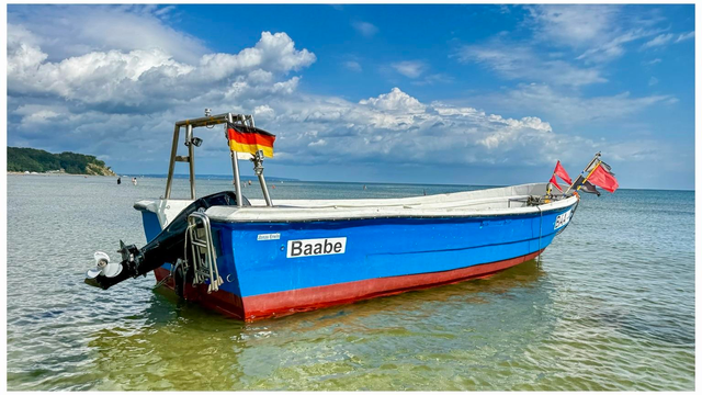 A blue fishing boat named "Baabe" is anchored in shallow water in the Baltic Sea, with a German flag and red flags waving above. The background features a clear sky with scattered clouds and a shoreline in the distance.