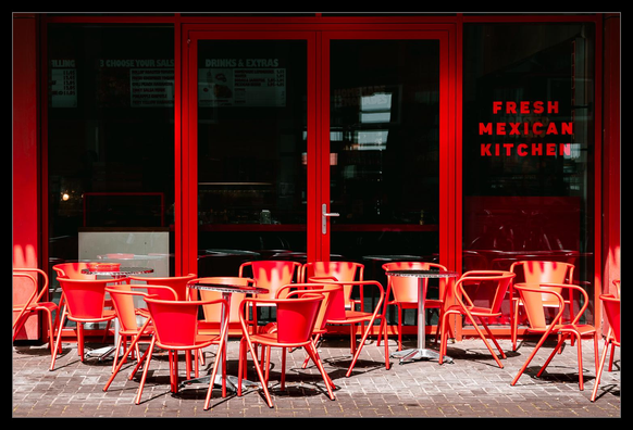 Eingangsbereich eines Restaurants. Wir blicken frontal auf eine Fensterfront mit Eingangstür und etwas unkoordiniert herumstehende Tische und Stühle. Die besonderheit ist, dass Tische, Stühle, Tür und Fensterrahmen in leuchtend rot sind.