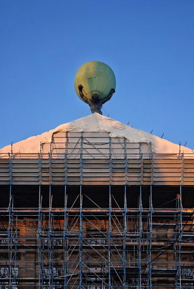 A photo of the top of a large building covered in scaffolding. At the top is a dark statue holding up a large green spherical object. The sky in the background is clear and blue.