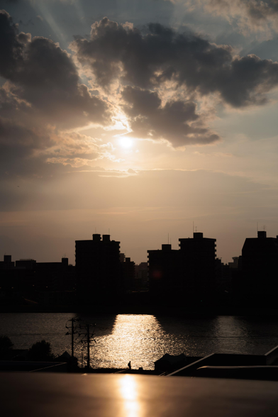 Silhouetted city skyline at sunset with clouds and sunlight reflecting on a river.