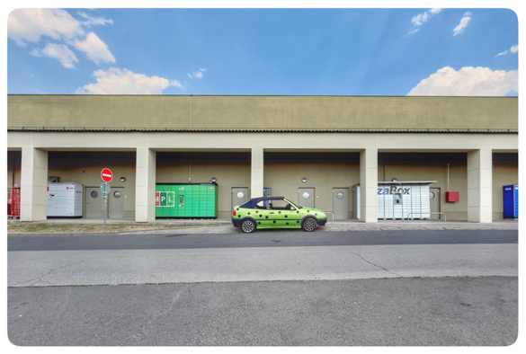 Ein grünes Auto mit schwarzen Punkten steht an einer Art überdachten Gang vor einem Gebäude. Der Bereich ist durch Pfeiler abgeteilt und in einigen davon stehen die Boxenwände unterschiedlich gefärbter Paketdienstleister.
