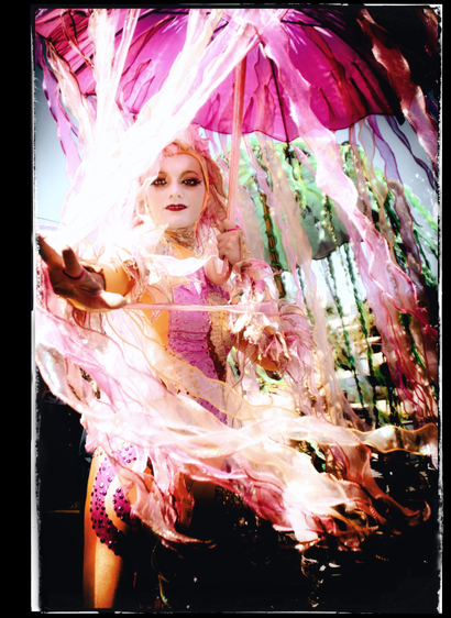 Street portrait.
The Castro Street Fair, Sunday, October 7th 2012. 

A picture of a woman with white mime face paint, dressed in a tight pink and gold jester outfit. She is standing in the shade of a pink parasol with dozens of long metallic ribbons... and she reaches out to me, waving one arm while casting a magical spell. My luck, it makes me extra gay.