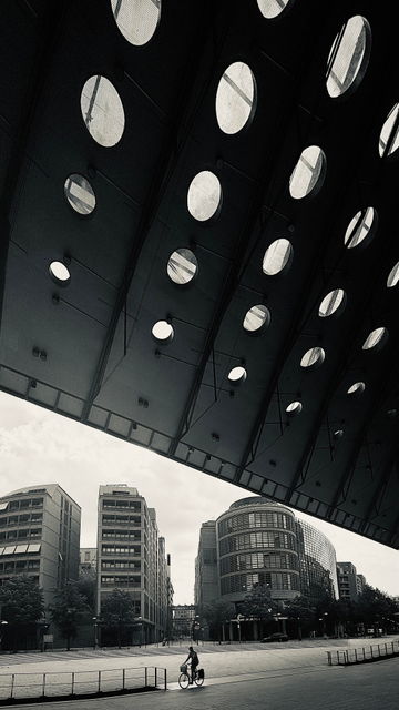 A cyclist rides through an empty urban area beneath a distinctive, perforated roof. Surrounding buildings feature modern architecture with glass and brick facades, and the scene is in black and white, giving it a moody atmosphere.