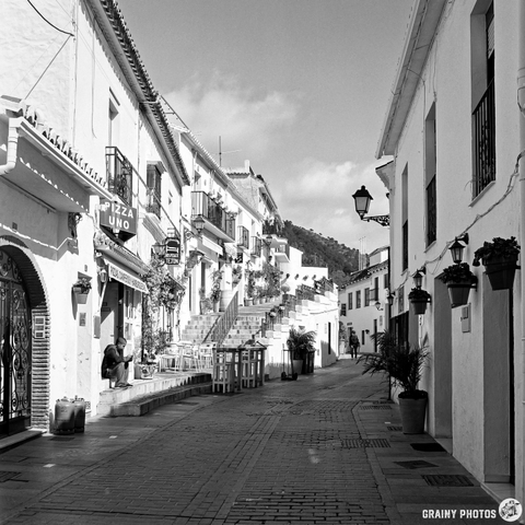 A charming cobblestone street lined with whitewashed buildings, featuring a restaurant offering pizza. Lush greenery in pots and a few people walking along the path create a serene atmosphere in a quaint, picturesque setting.