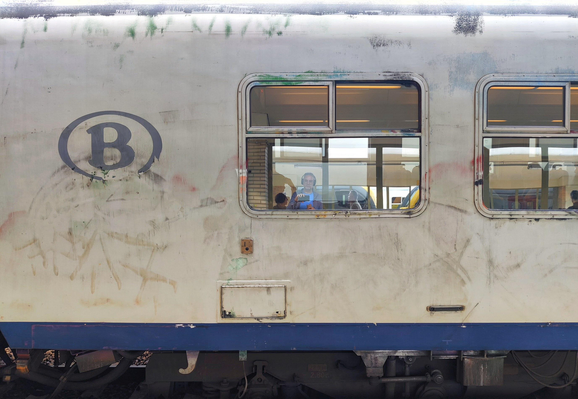 A close-up photo of a silver blue train car. The side of the train features a large blue circle with a "B" logo on a surface that shows signs of wear (and/or imperfect grafitti removal?). The windows of the train are slightly open, and  people on the platform are reflected in the glass.