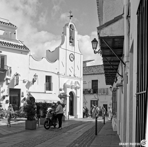 A black and white street scene in Mijas, featuring traditional white buildings, Iglesia de San Sebastián with a clock, and people strolling. Potted plants adorn the area, and there are lanterns hanging from the buildings, capturing a charming atmosphere.