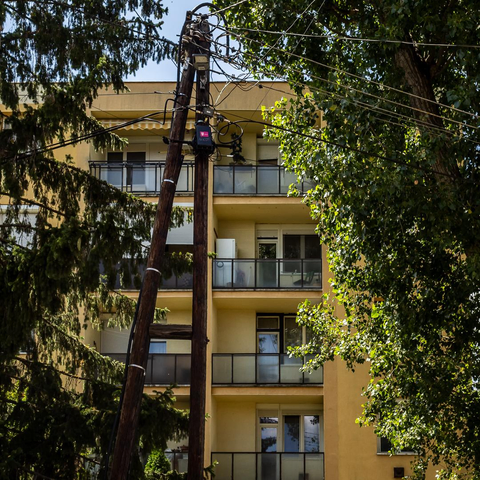Zwei schräg stehende Holzmasten mit zahlreichen Kabeln vor einem gelben Mehrfamilienhaus. Zwischen den Bäumen sind mehrere Balkone und Fenster zu sehen.