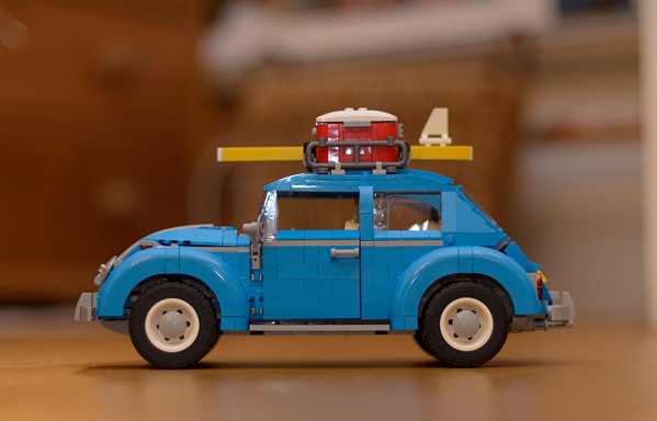 A photo of a blue LEGO Volkswagen Beetle car with a suitcase and surfboard mounted on the roof. The model stands on a wooden surface, with a blurred background.