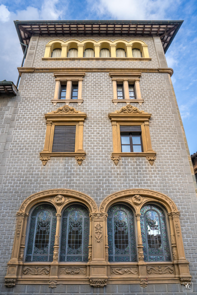 Color photo looking up the facade of a multi-story building tower that has an eclectic mix of classical, baroque, and modernisme elements including elaborately decorated stained glass windows on the lower level.