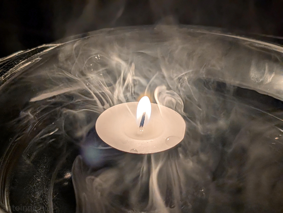 A close-up of a lit floating tea light candle inside a round glass bowl filled with swirling white smoke against a dark background.

The classic white disc tea light candle floats at the center. Its bright flame emits a warm, bright glow at the core with a visible wick. 

Thick, ethereal wisps of white smoke swirl dynamically around the floating candle.